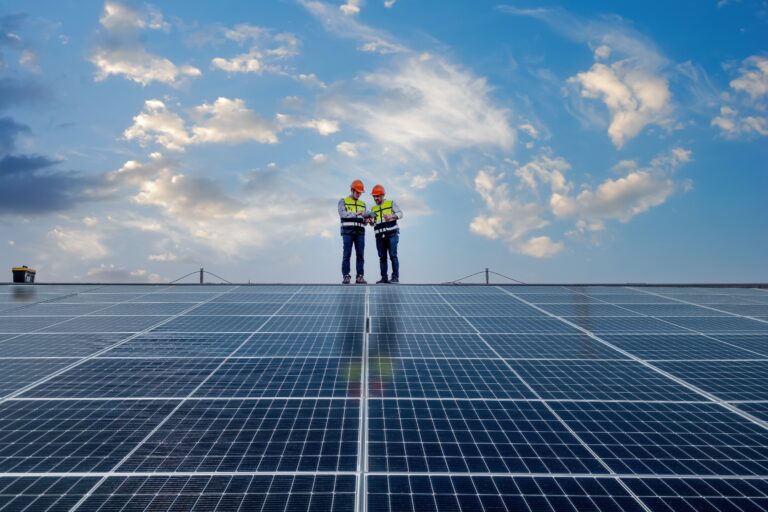 Blog 1 Zwei Arbeiter in Sicherheitsausrüstung stehen vor einem bewölkten blauen Himmel auf einer Solaranlagen-Anlage.