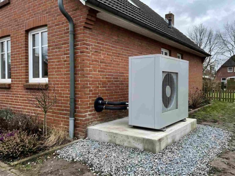 Neu aufgestelltes Wärmepumpen-Außengerät auf einem Betonsockel mit Kiesbett vor einer Klinkerfassade, gedämmte Leitungen führen durch die Wand