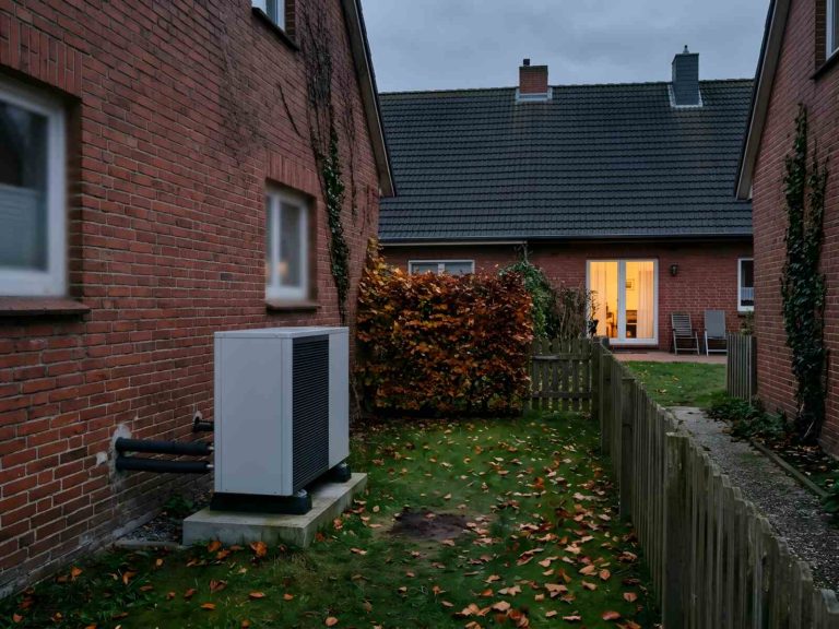 Außengerät einer Wärmepumpe im schmalen Garten zwischen zwei Backstein-Reihenhäusern, davor eine niedrige Hecke