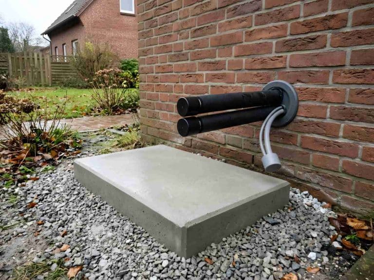 Leerer Betonsockel mit Kiesbett vor einer Klinkerwand, aus der Wanddurchführung ragen gedämmte Leitungen und ein Elektrokabel