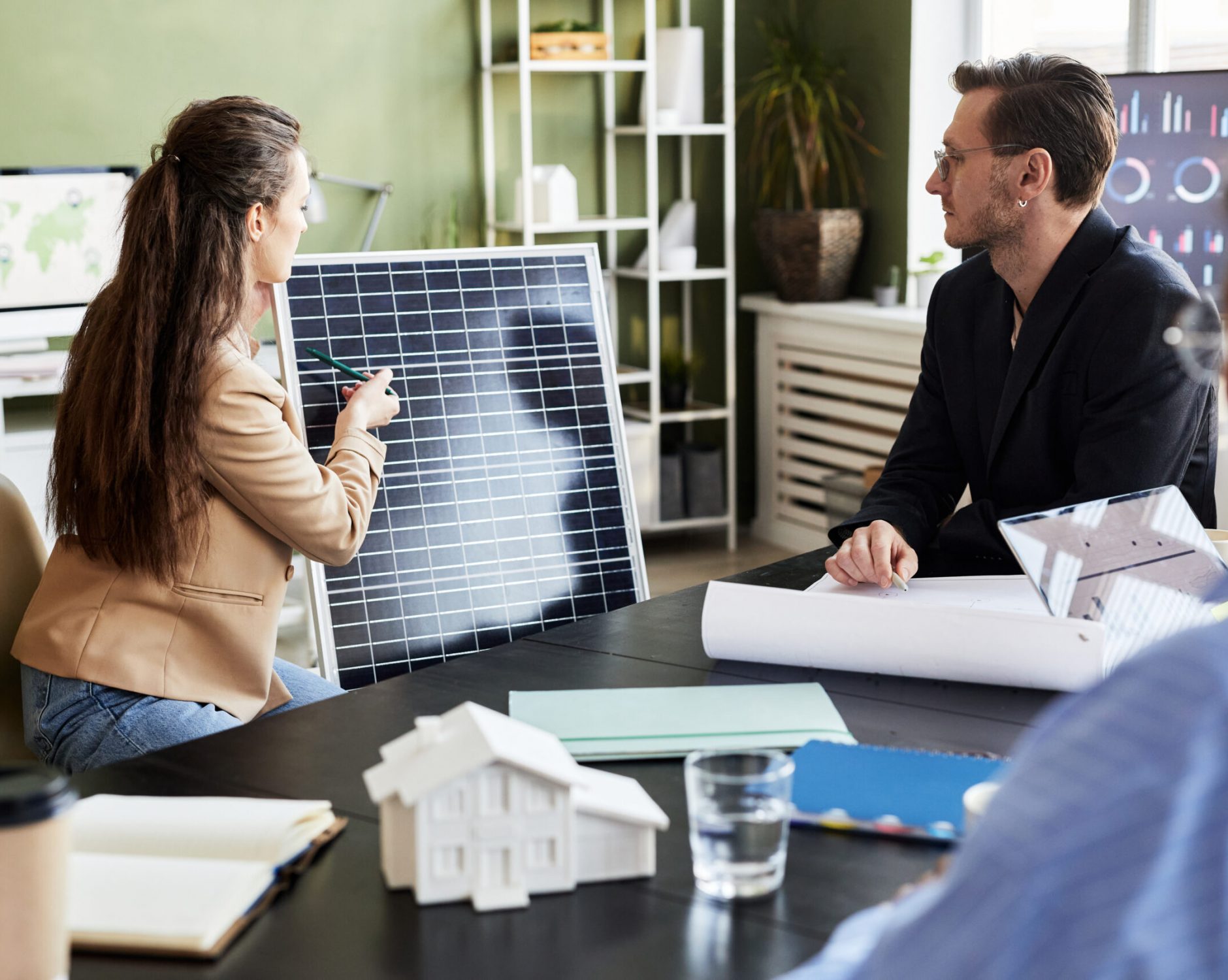 Eine Frau präsentiert zwei Kollegen in einem Büro ein Solarmodul und betont dessen photovoltaische Effizienz. Ein Modellhaus und Baupläne auf dem Tisch veranschaulichen ihr nachhaltiges Energieprojekt.